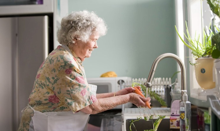 Elderly woman washing vegetables in a bright kitchen.